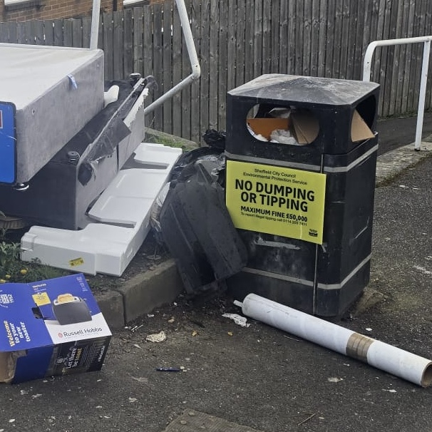Photo of a bin with a large sticker saying "no dumping or tipping" surrounded by large dumped materials.